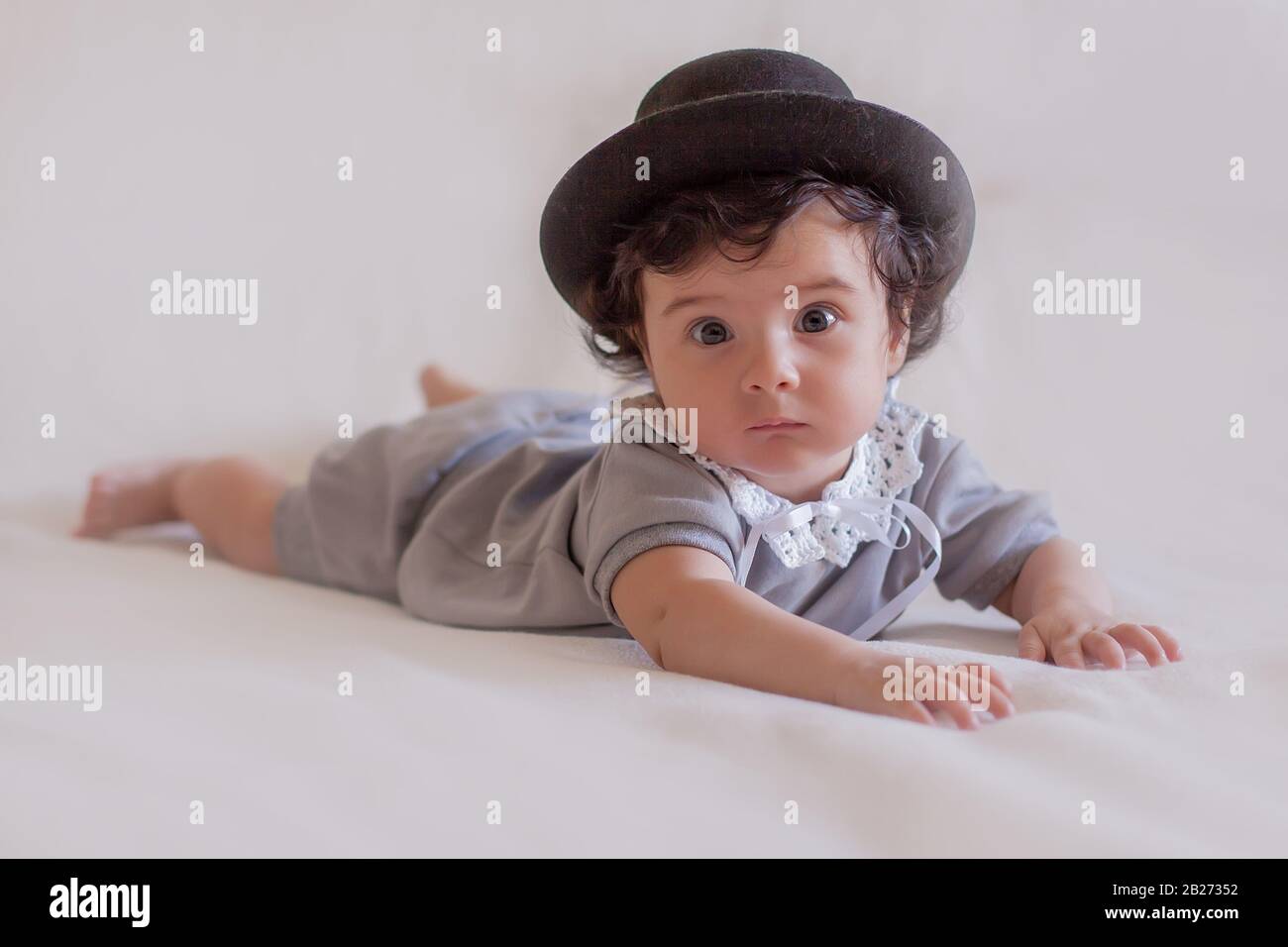 Un joli petit bébé dans un chapeau noir et un costume gris vintage et un col en dentelle blanc se trouvent sur un plaid blanc. Banque D'Images