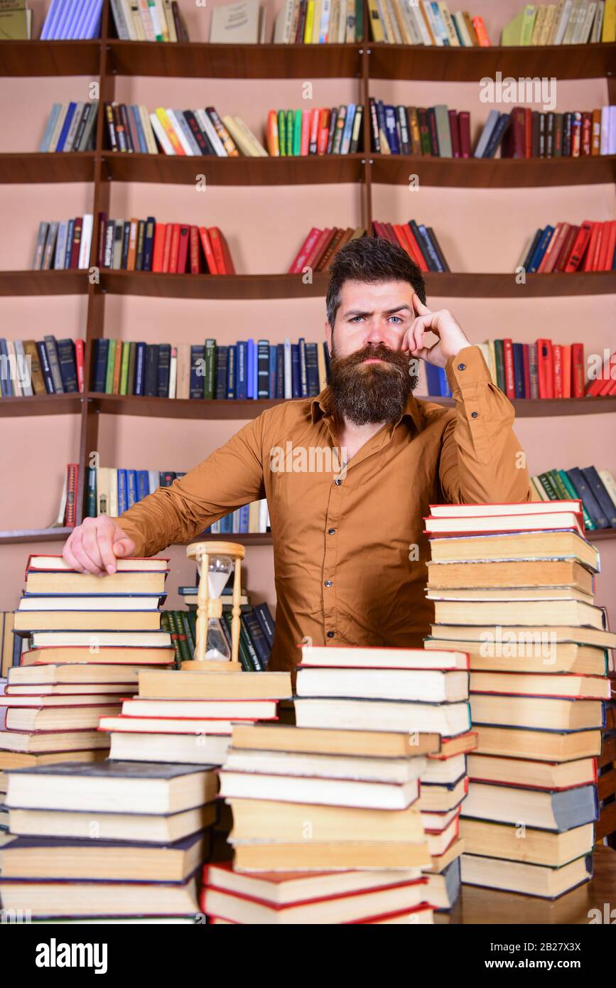 Professeur ou étudiant avec des pieds de barbe à la table avec des livres, défocused. Concept de bibliothécaire. L'homme sur le visage réfléchi se tient entre des piles de livres, tout en étudiant dans la bibliothèque, les étagères de livres sur l'arrière-plan Banque D'Images