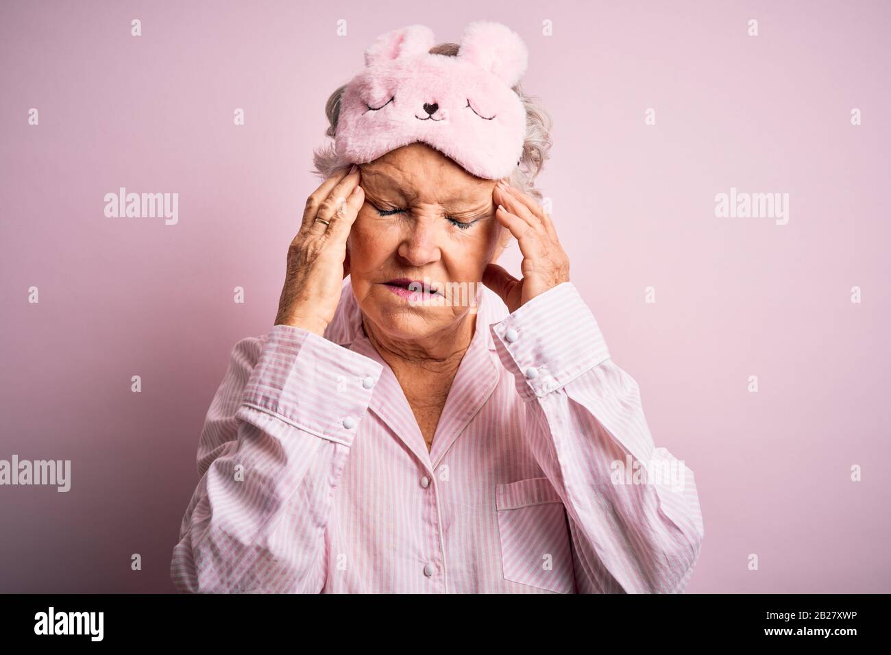 Senior belle femme portant masque de sommeil et de pyjama sur fond rose isolé avec main sur la tête pour la douleur dans la tête parce que le stress. Souffrance migrai Banque D'Images
