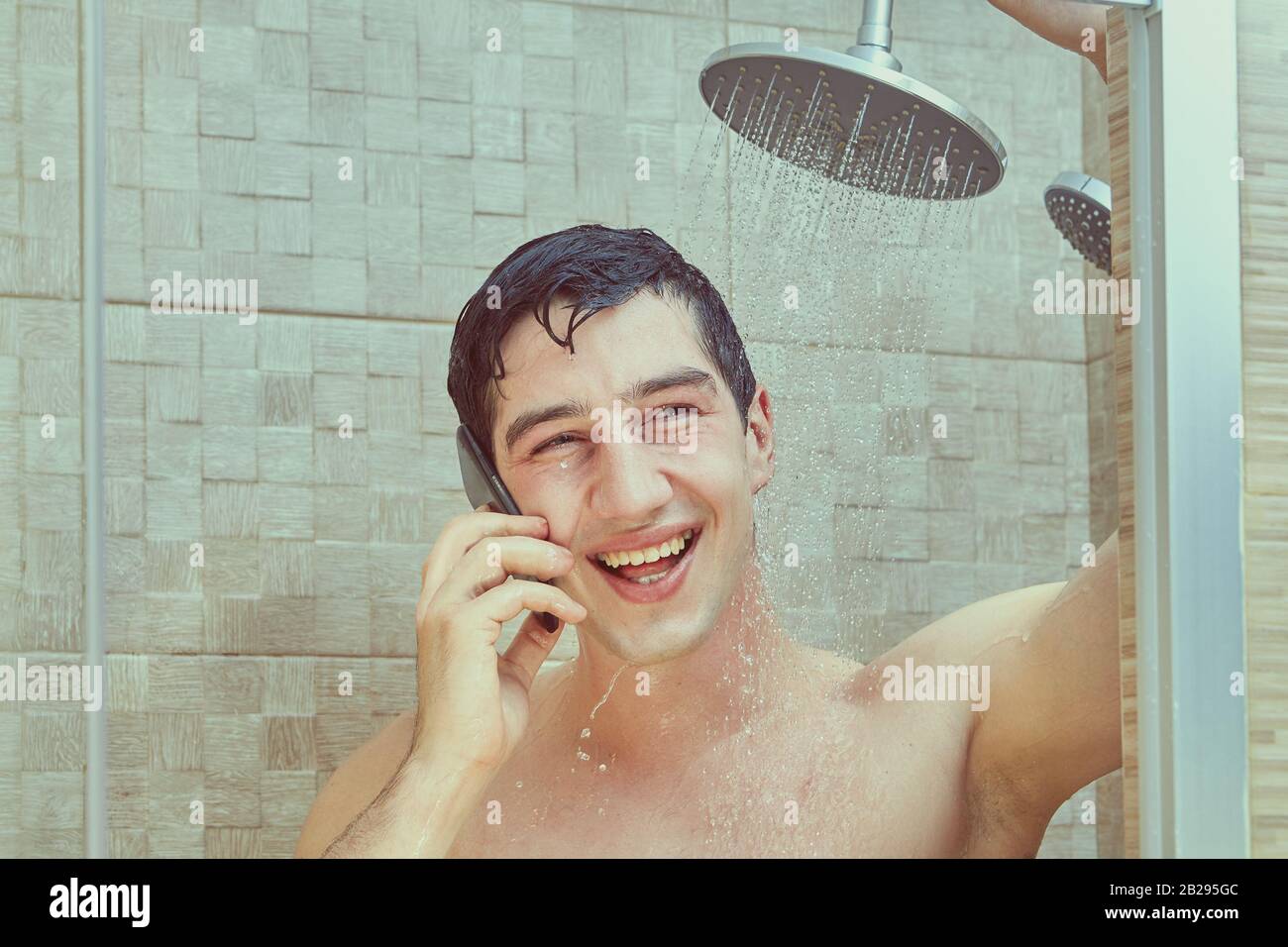 Rire l'homme blanc utilise un smartphone, debout sous les ruisseaux de l'eau qui coule de la douche. Un jeune homme caucasien parle au téléphone dans le b Banque D'Images