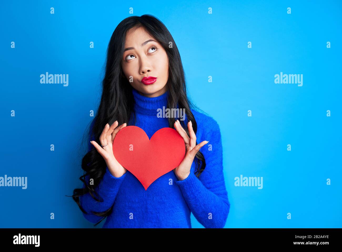 Jolie jeune femme tenant son cœur en papier dans les mains et regardant isolé sur fond bleu. L'amour et le concept romantique Banque D'Images