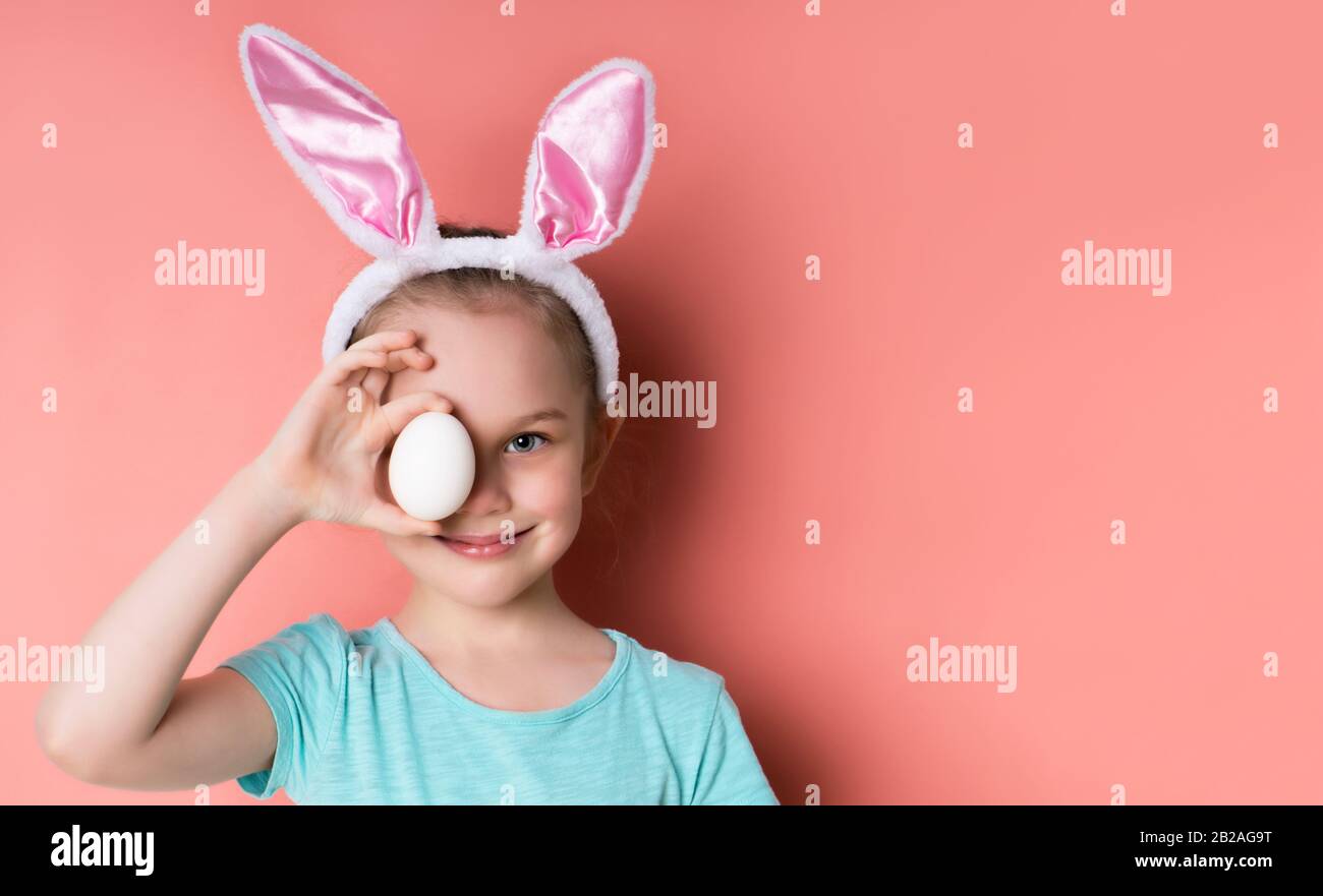 Petite fille dans le bandeau avec oreilles de lapin, t-shirt bleu. Elle  souriait, couvrait ses yeux avec de l'oeuf, posant sur fond rose. Pâques  Photo Stock - Alamy