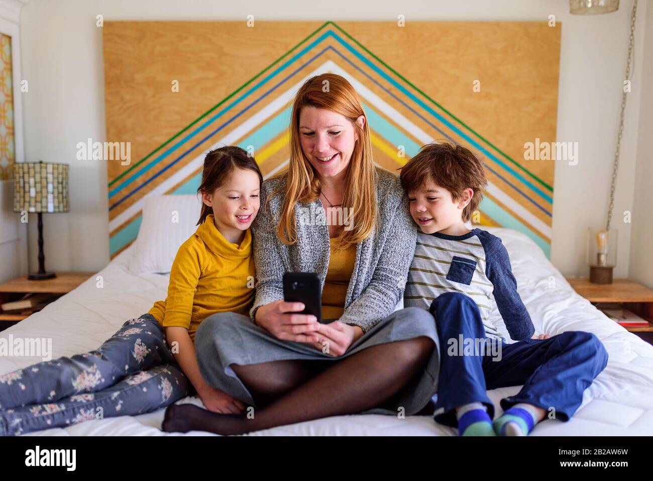 Mère assise au lit avec deux enfants regardant un téléphone mobile Banque D'Images