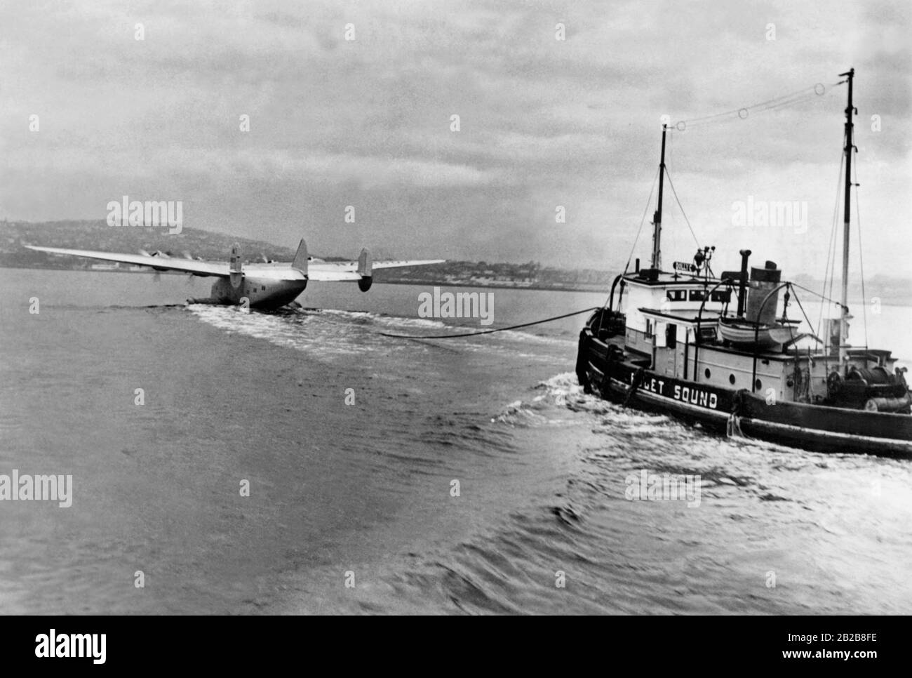Un Boeing 314 « Clipper » en bateau-pilote pendant les vols d'essai sur le Puget Sound dans l'État de Washington. Le bateau volant tire un remorqueur dont le moteur tourne vers l'arrière à pleine puissance. Le remorqueur empêche le bateau volant de se retirer. Banque D'Images