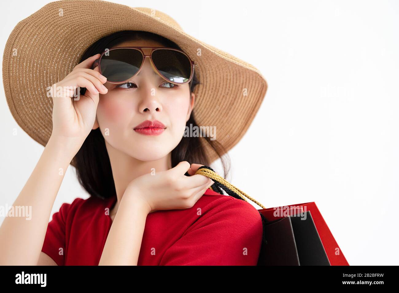 Portrait d'une femme asiatique portant une robe rouge et un chapeau d'été portant des sacs à provisions et un sourire heureux sur fond blanc. Banque D'Images