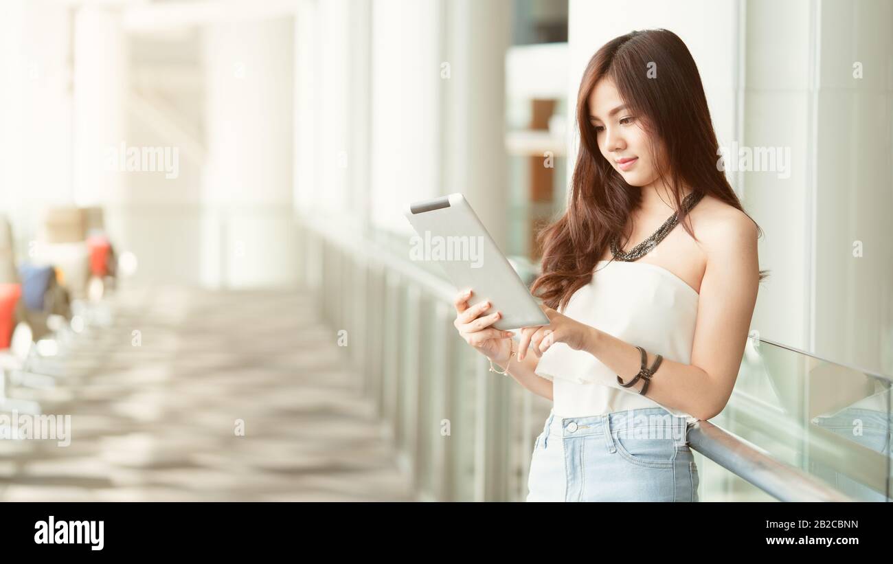 Jeune femme asiatique belle travaillant et jouant sur sa tablette tout en se tenant facilement à côté du couloir, concept travaillant sur la social n'importe où, backg Banque D'Images