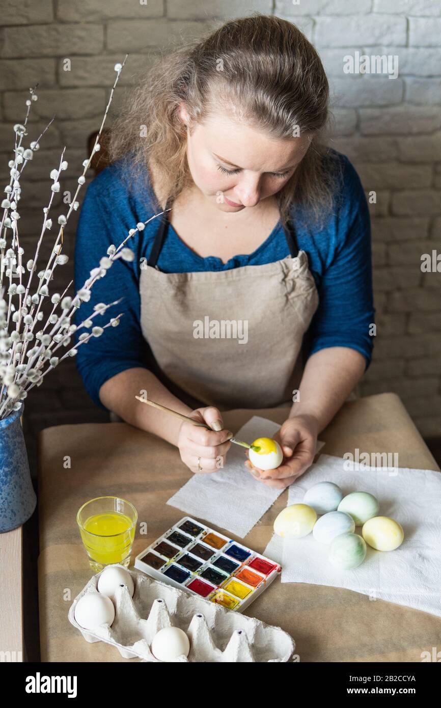 Jeune femme blonde peignant des œufs aux aquarelles pour la célébration de Pâques. Vase avec des chatons au premier plan Banque D'Images