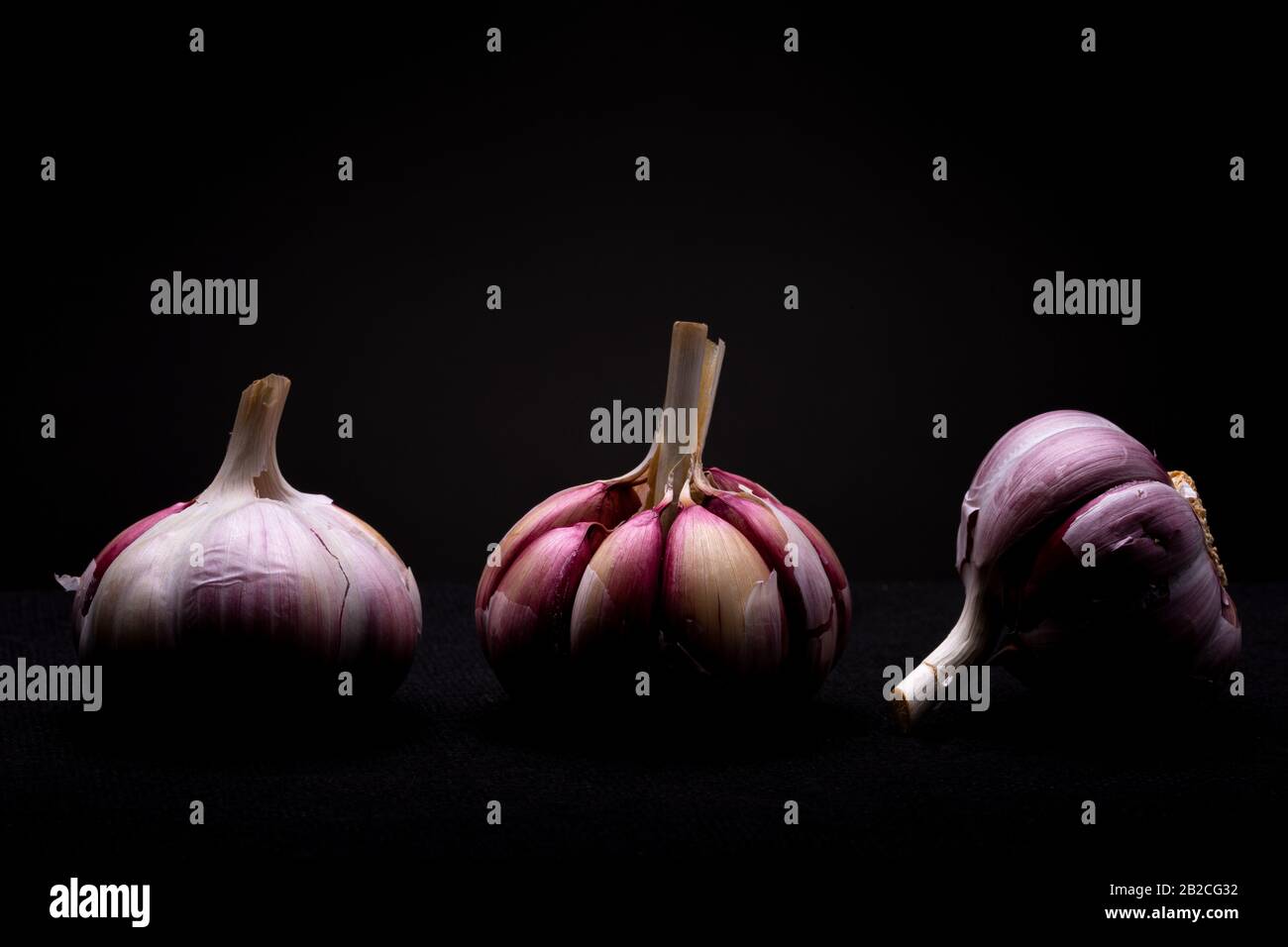 Gros plan sur trois lots d'ail rouge avec la peau pelée du milieu montrant les pièces individuelles en studio avec une lumière dramatique sur un fond sombre Banque D'Images