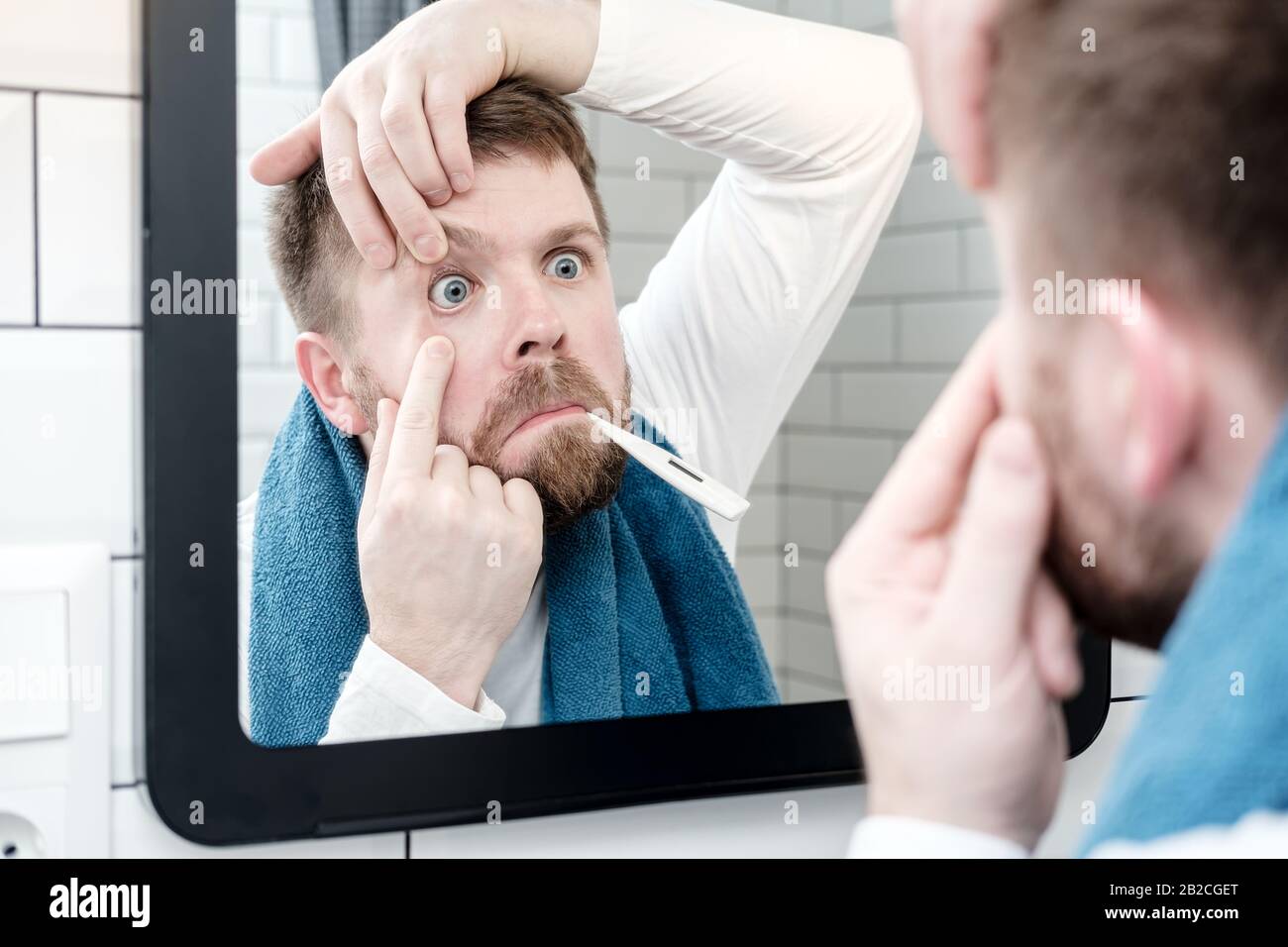 Un homme effrayé avec un thermomètre dans la bouche regarde dans le miroir et examine son oeil dans la salle de bains. Concept de santé. Banque D'Images