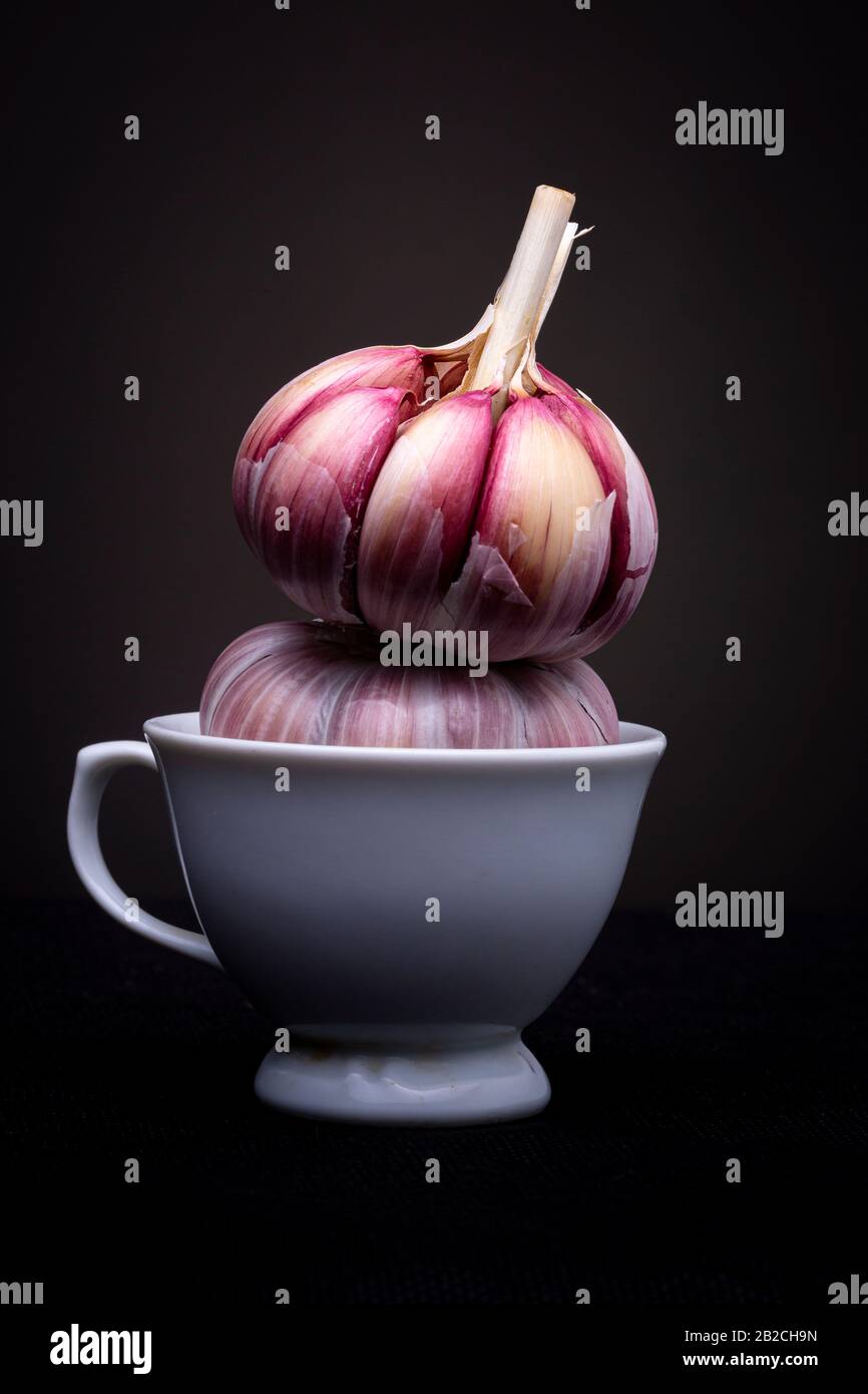 Ail rouge ou violet empilé dans une petite tasse blanche dans un cadre studio avec un fond clair et sombre dramatique Banque D'Images