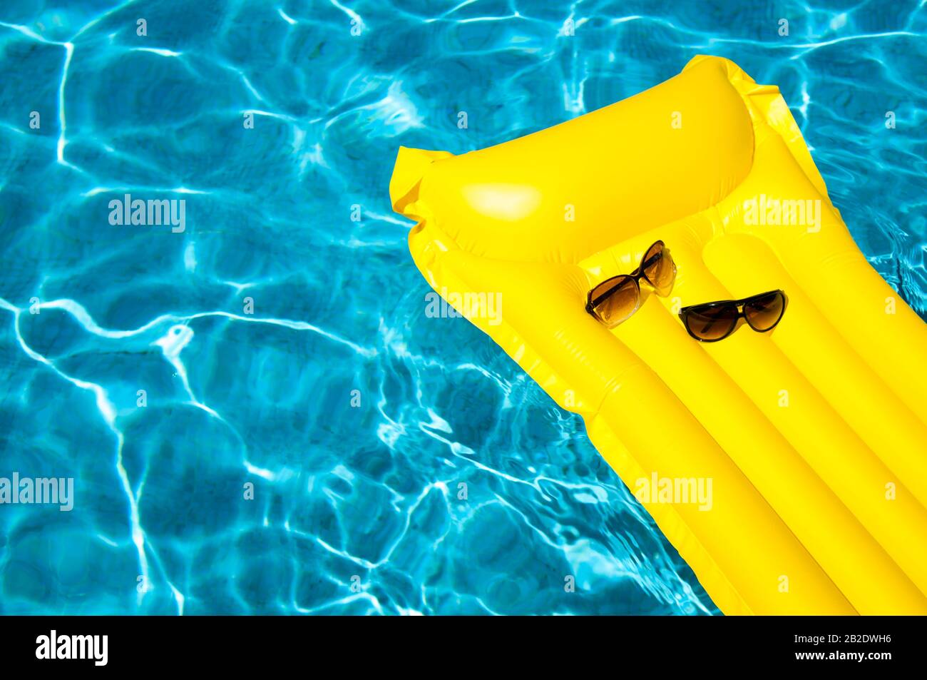 Ses lunettes de soleil et ses hers reposent sur un radeau gonflable jaune vide flottant dans une piscine bleu vif Banque D'Images