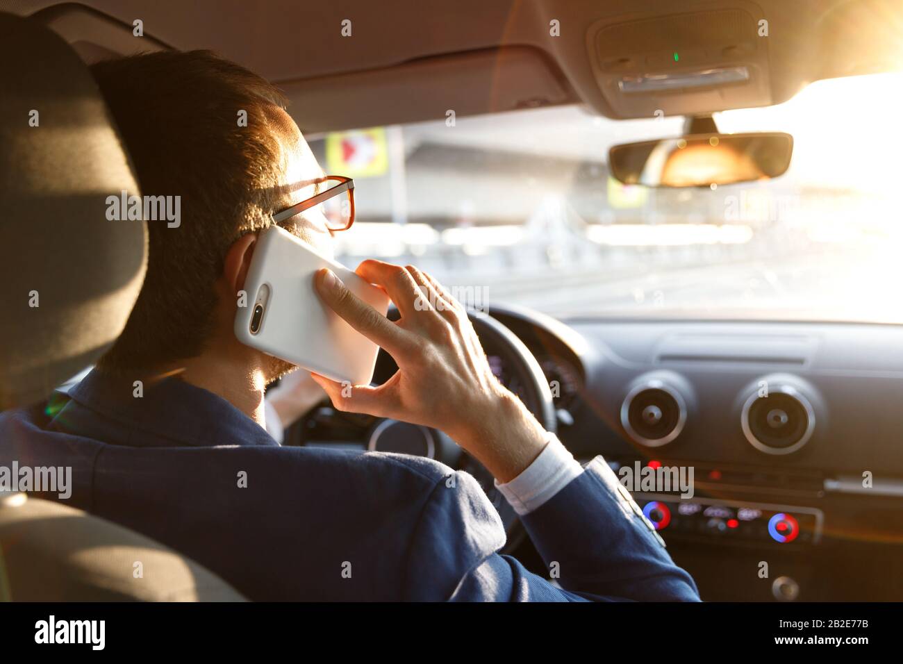 Homme en blazer en utilisant et en parlant sur smartphone tout en conduisant une voiture au coucher du soleil, en regardant attentivement la route, vue arrière. Banque D'Images