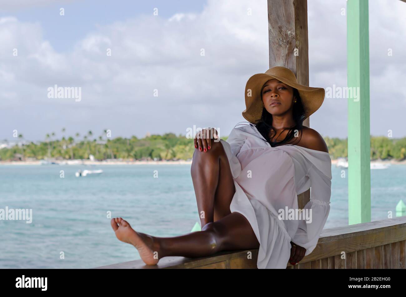 Femme noire de 25 à 30 ans, assise au port, vêtue de modèles blancs, de vêtements frais d'été et tropicaux, avec paysage de plage et ciel ensoleillé Banque D'Images