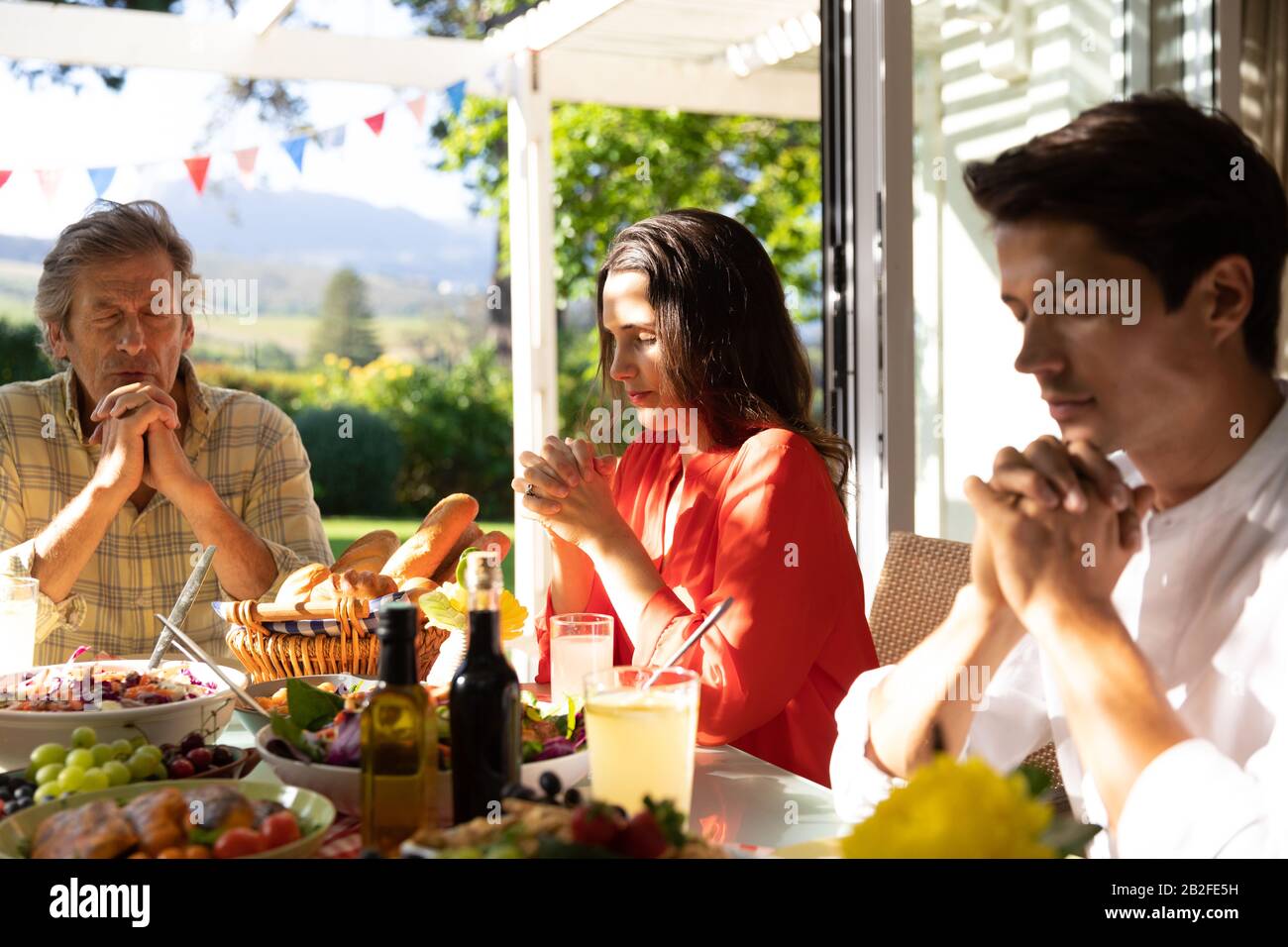 Vue de face d'un homme caucasien et de sa fille et de son fils adultes assis dehors à une table de dîner pour un repas, avec des mains claquées et des yeux fermés dans p Banque D'Images