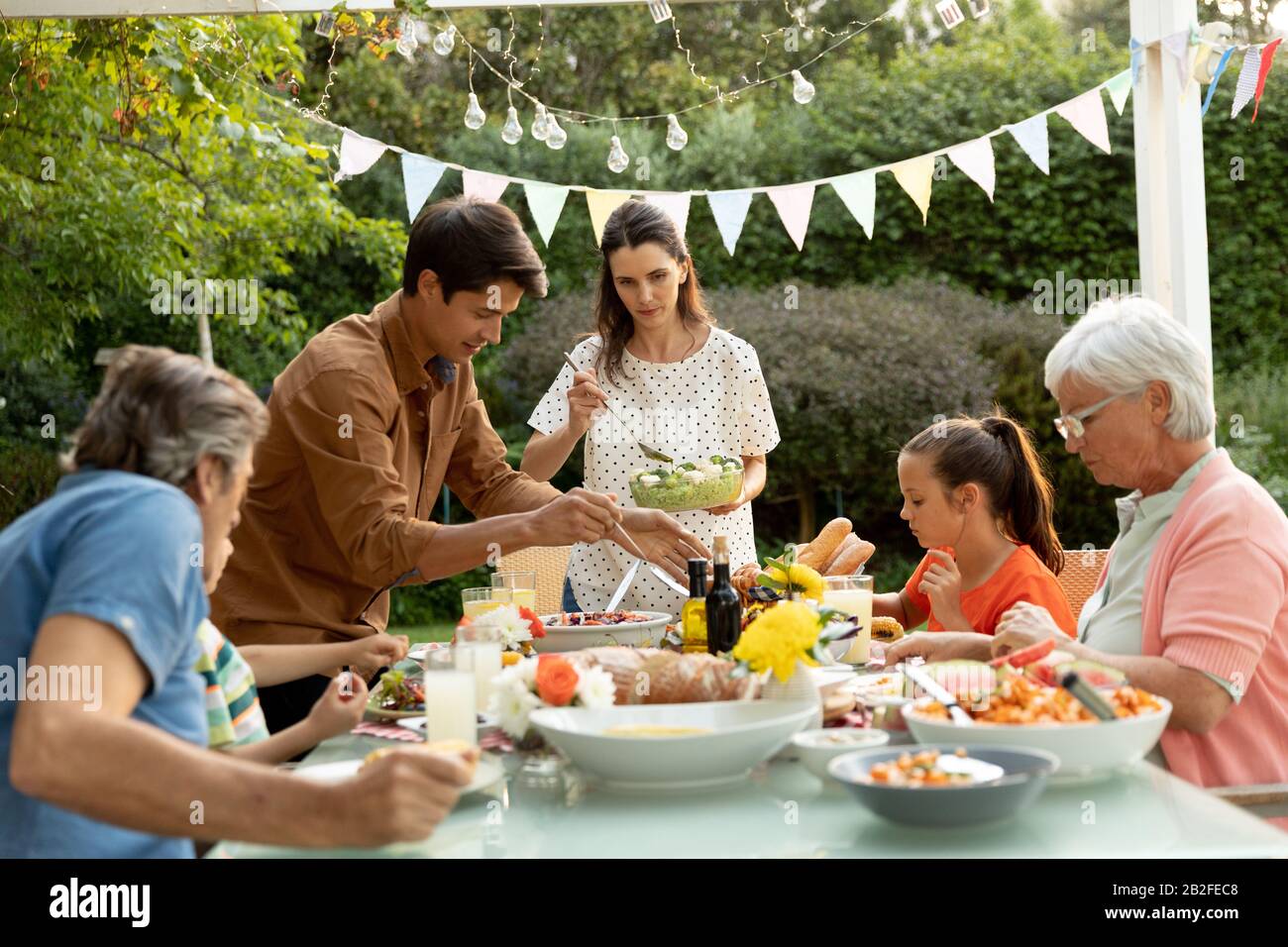 Vue de face d'une famille caucasienne de plusieurs générations assise à l'extérieur d'une table de dîner pour un repas, servant les autres plats. Famille profitant du temps à h Banque D'Images