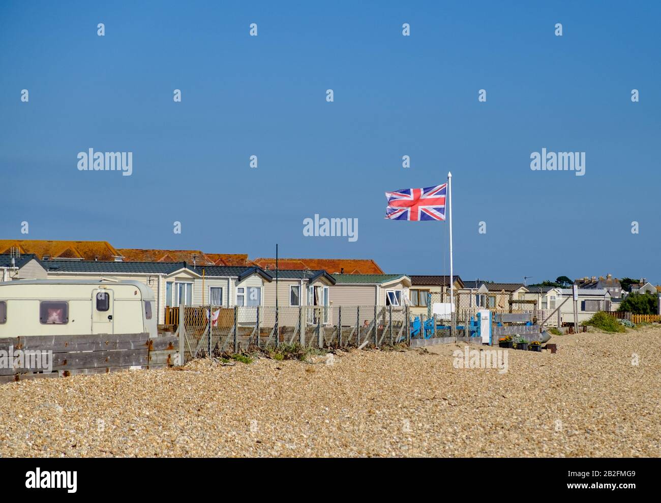 Caravanes blanches statiques derrière une clôture avec plage de galets et drapeau britannique sur le poteau au West Sands Caravan Park, Bunn Leisure, Selsey, West Sussex, Angleterre. Banque D'Images