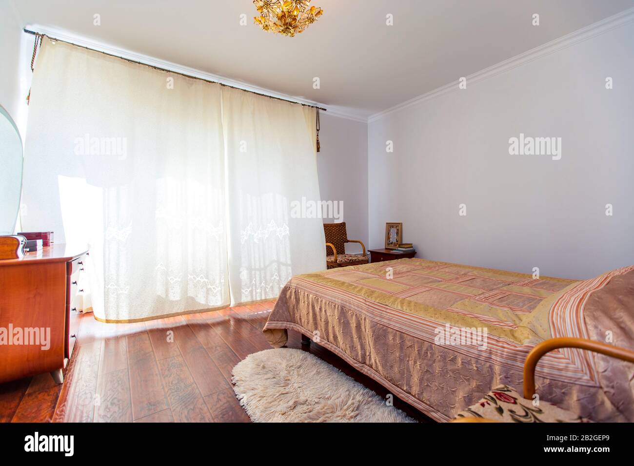 Une grande chambre avec un lit en acajou, des chaises en osier et une élégante coiffeuse avec miroir semi-circulaire. Le lit a une couverture beige en carrés. Banque D'Images