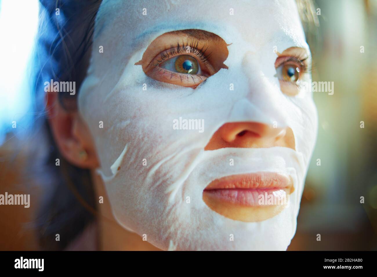 Portrait d'une femme de ménage élégante de 40 ans en pyjama avec masque facial en feuille blanche sur le visage dans la maison moderne en hiver ensoleillé. Banque D'Images