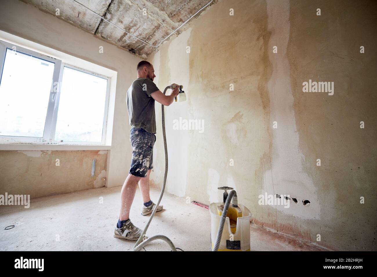 Homme peint un mur gris avec pistolet à vaporiser, rénover de la nouvelle maison. Ouvrir le récipient avec une peinture. Vaporiser de la peinture. Le mâle est vêtu de vêtements enduits de peinture. Petite chambre avec fenêtre et lumière du jour Banque D'Images