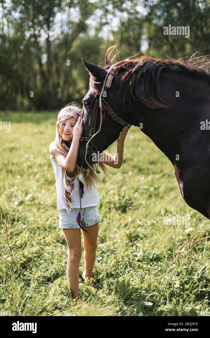Belle fille caucasienne heureuse portant des vêtements décontractés et des accessoires de boho dans les cheveux, souriant et reposant sa tête sur le cheval au soleil, à l'extérieur Banque D'Images