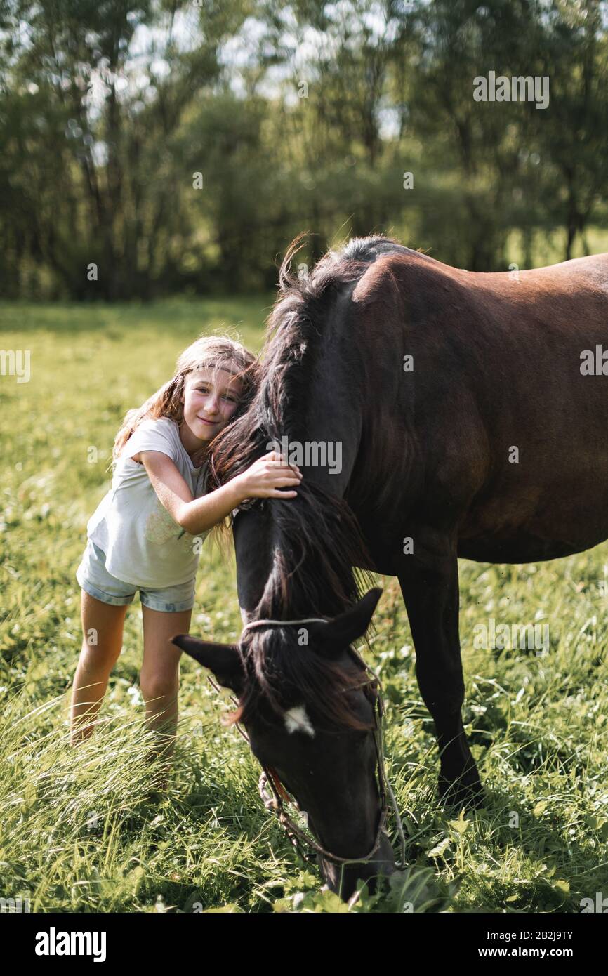 Petite fille en t-shirt blanc et pantalon de Jean embrassant et paetant un cheval sur une journée ensoleillée et vive à l'extérieur dans le champ avec de l'herbe verte Banque D'Images