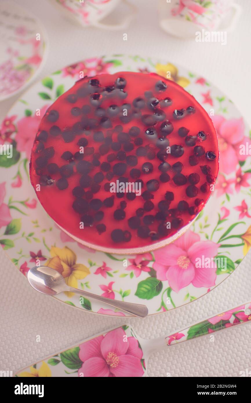 Cheesecake à la cassis dans un plat à fleurs. Cuillère, spatule à pâtisserie, tasses pour thé et soucoupes sur une table blanche. Banque D'Images