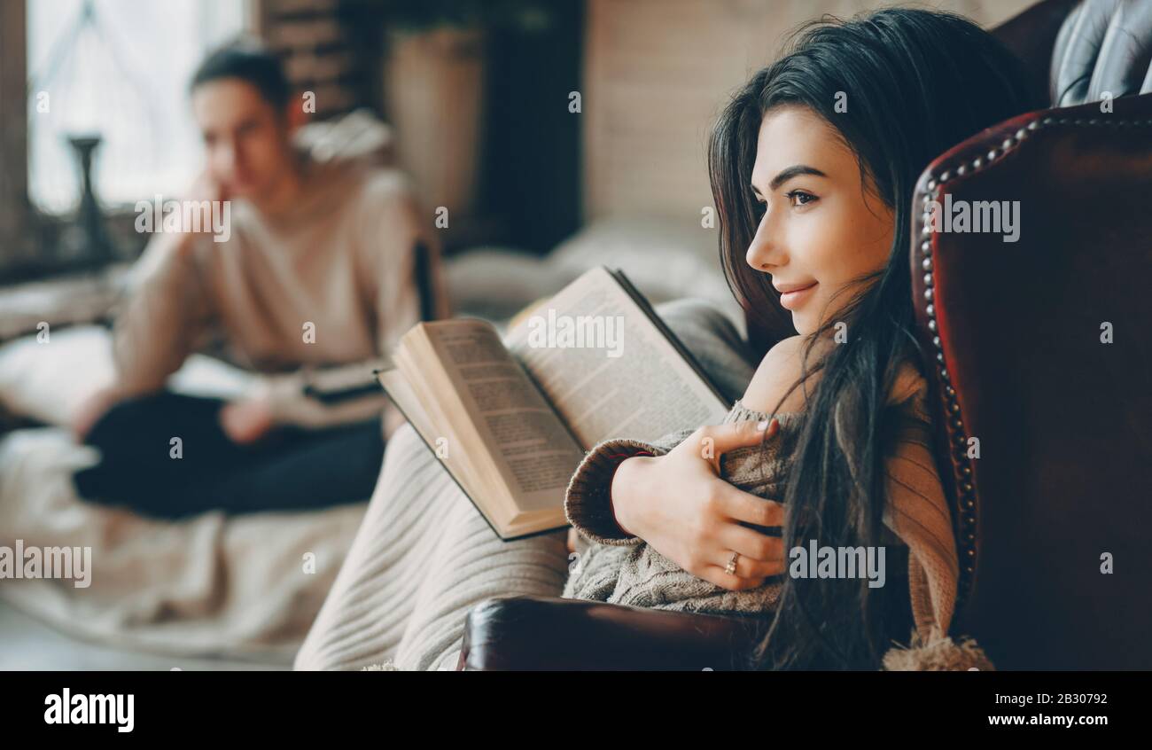 Une merveilleuse fille caucasienne aux cheveux noirs lit un livre alors que son amant est assis sur le canapé et la regarde Banque D'Images