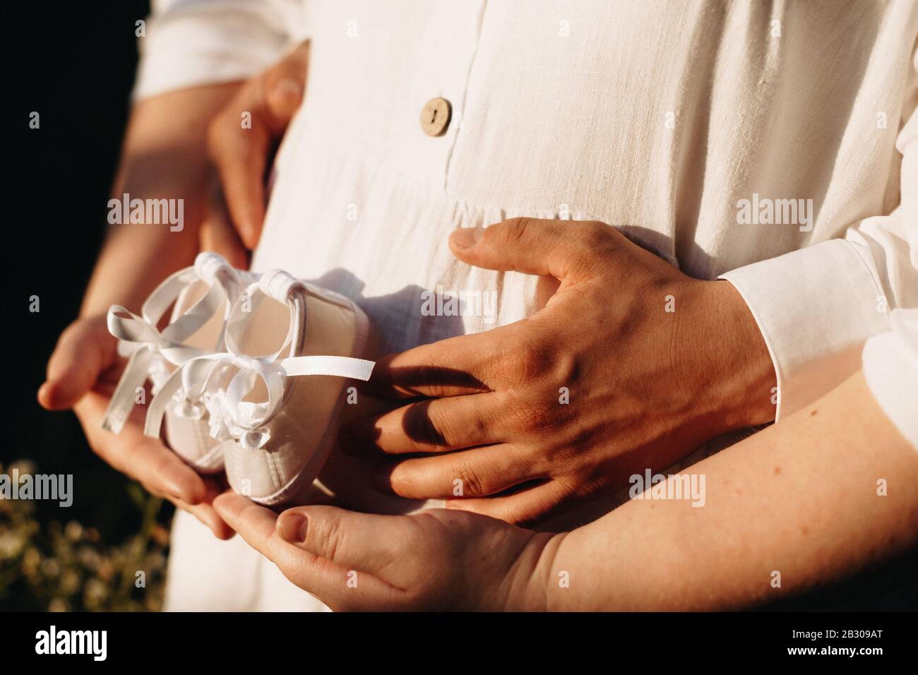 Charmante femme enceinte embrassée par son mari tout en posant à la lumière du soleil avec une paire de chaussures de bébé Banque D'Images