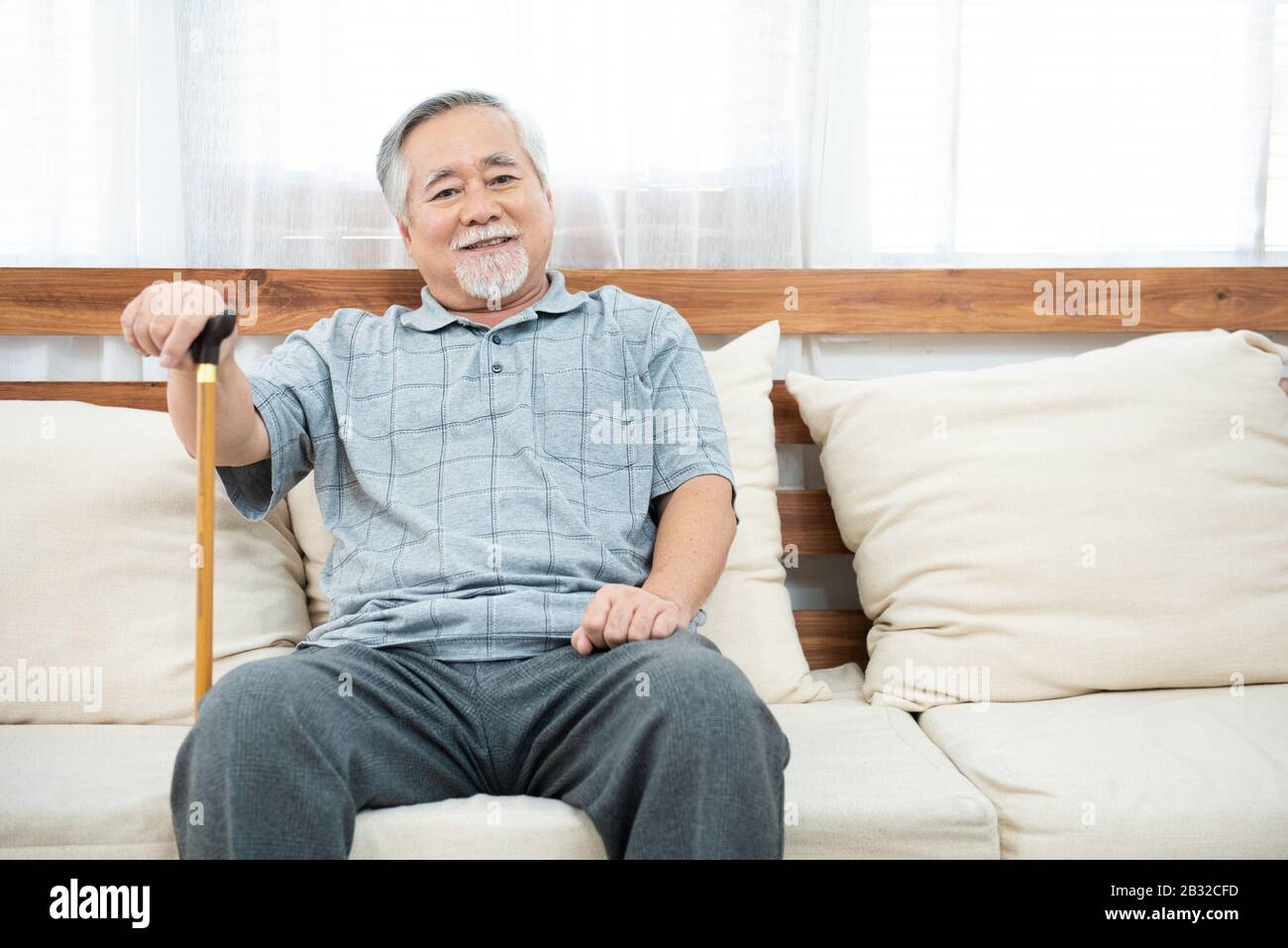 vieux homme âgé assis, reposant ses mains sur un bâton de marche en bois assis sur le canapé dans la salle de séjour de la maison après la retraite. Banque D'Images