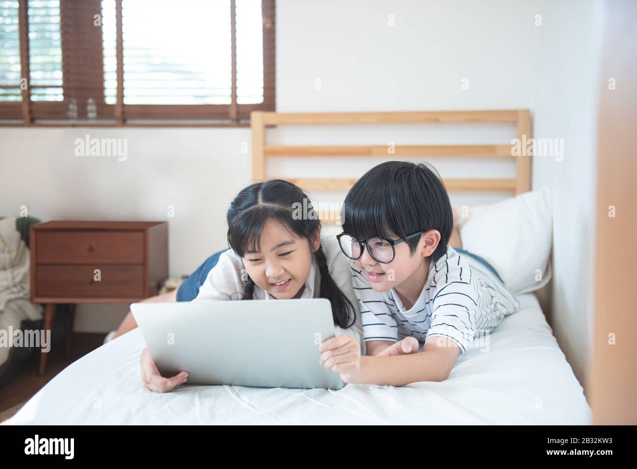 Joyeux asian garçon et fille jeu dans ordinateur portable allongé sur le lit dans la chambre à coucher dans la maison, frère et soeur utilisant ordinateur portable faire des devoirs, concept d'éducation. Banque D'Images