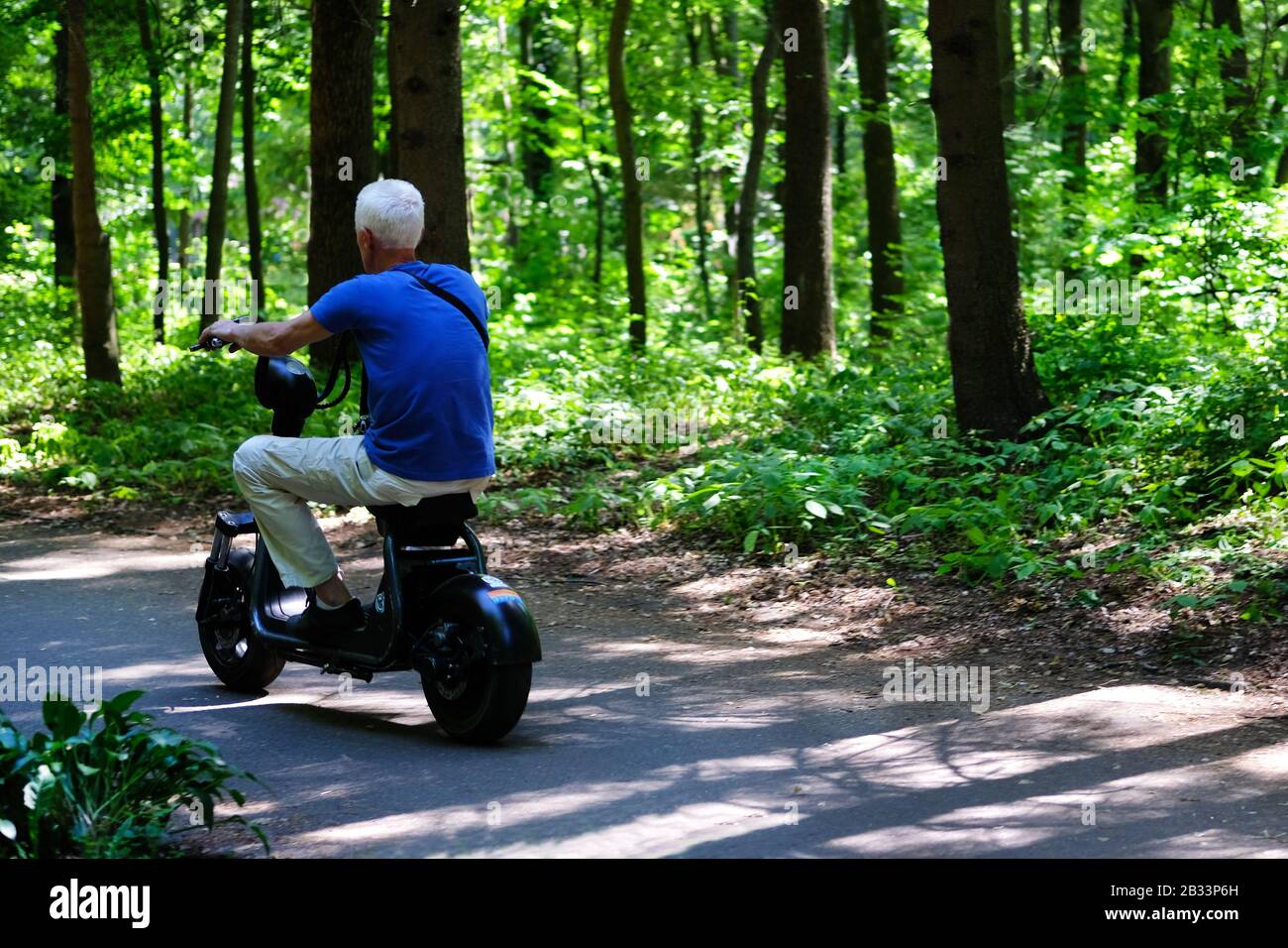 Homme senior en T-shirt bleu qui conduit de manière délice son scooter électrique parmi de nombreux arbres verts. Banque D'Images