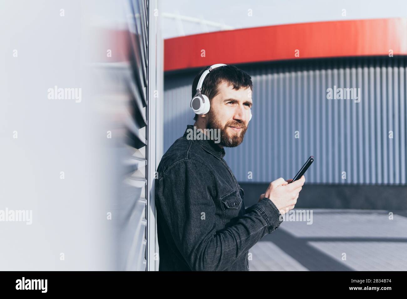 Jeune homme barbu écoutant de la musique dans des écouteurs sans fil dans l'environnement urbain, tenant le téléphone mobile dans ses mains Banque D'Images