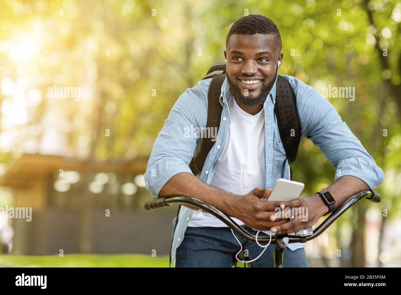 Joyeux Afro Man Assis Sur Vélo Et Écoutant De La Musique Sur Smartphone Banque D'Images