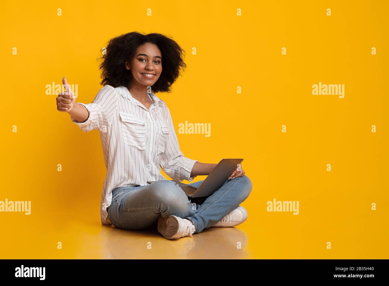 Afro Girl Souriant Assis Avec Un Ordinateur Portable Et Montrant Thumb Up Gesture Banque D'Images