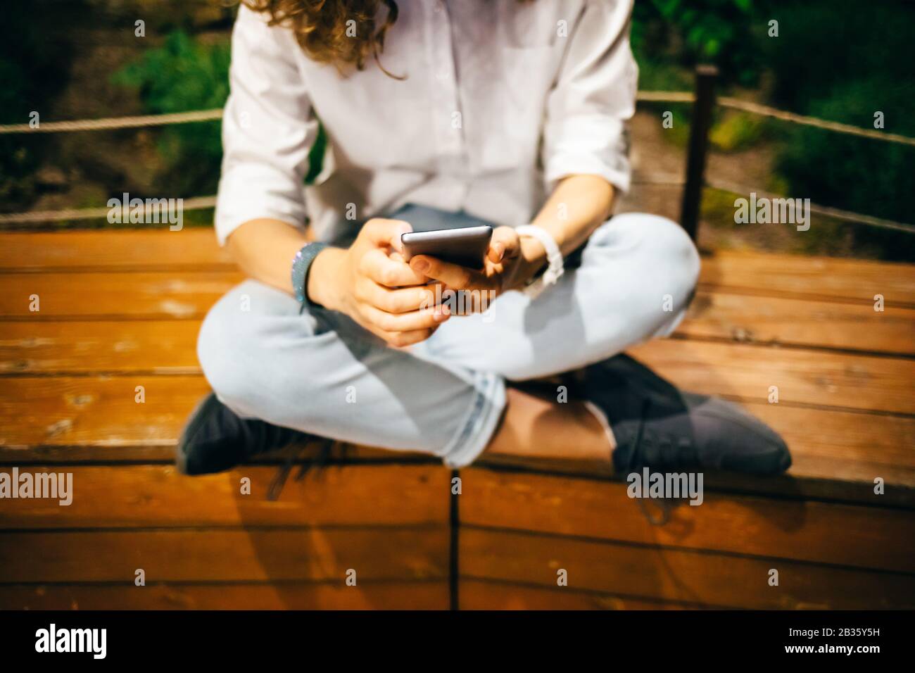 Jeune femme assise détendue sur un banc en bois avec ses jambes croisées tenant l'appareil mobile dans ses mains regardant l'écran. Fille utilisant smartphone été Banque D'Images