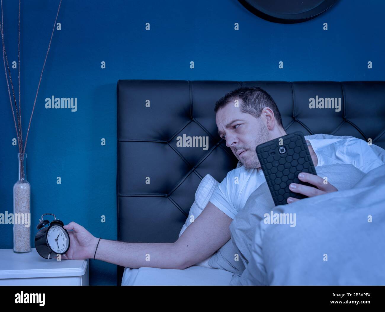un homme travaillant sur son smartphone la nuit regarde son réveil et ne peut pas dormir Banque D'Images