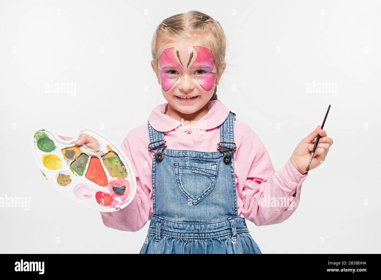 enfant souriant avec peinture aux papillons sur la palette de maintien du visage et le pinceau tout en regardant l'appareil photo isolé sur blanc Banque D'Images