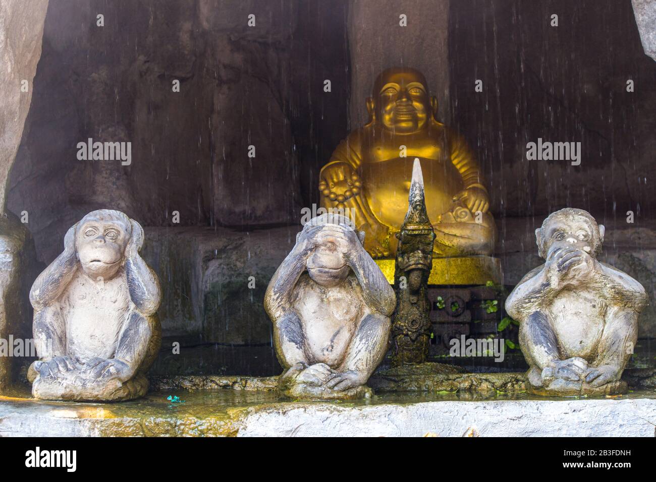 Bouddha et trois statues de singe s'assoient et ferment leurs oreilles, les yeux et la bouche dans une grotte avec chute d'eau sur fond flou. Banque D'Images