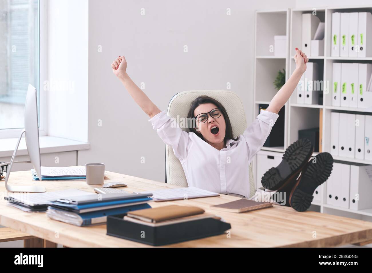 Une jeune femme fatiguée assise avec des pieds sur la table et bâillant avec des bras tendus au bureau Banque D'Images