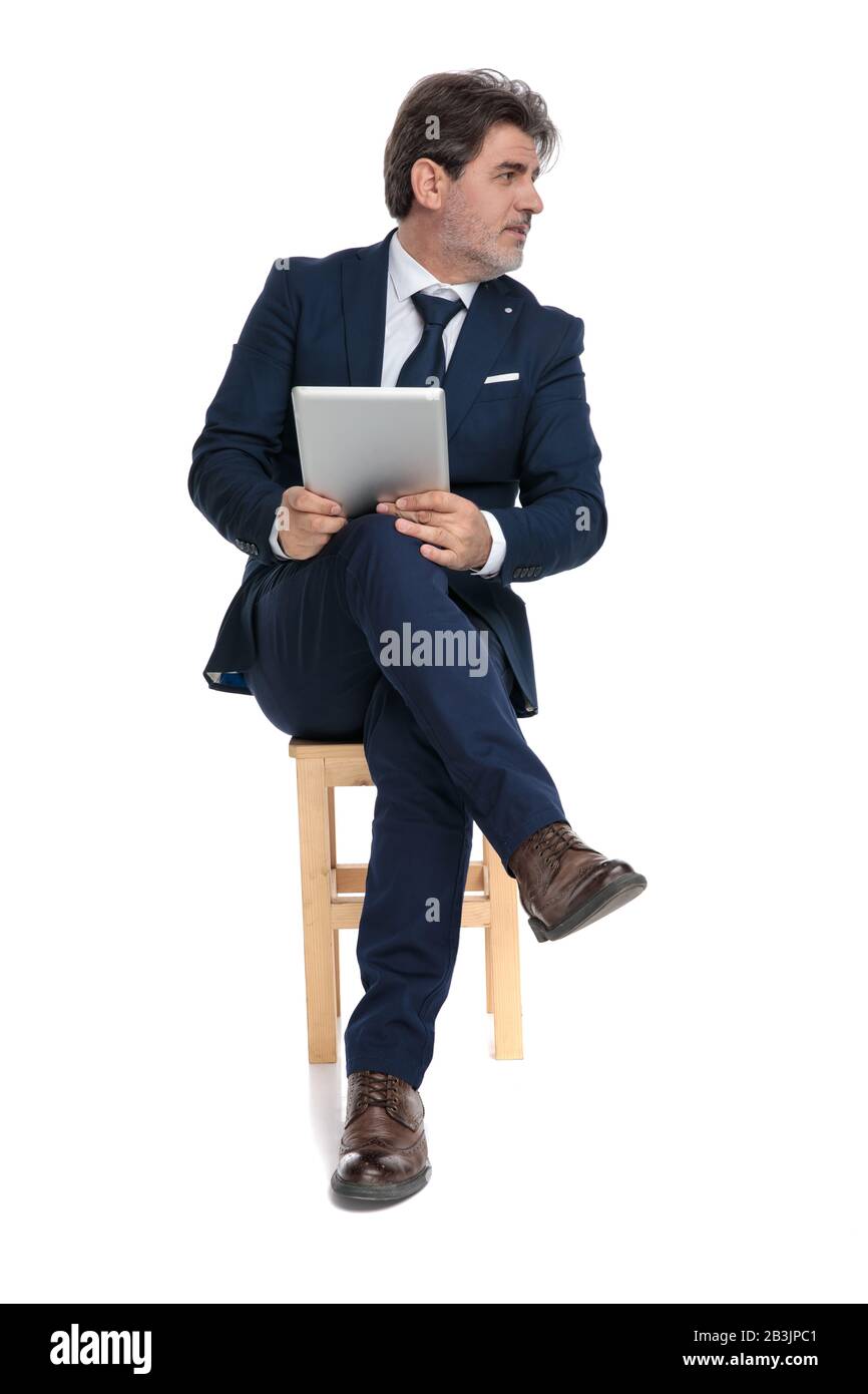un homme d'affaires raffiné et habillé avec un costume bleu marine est assis avec des jambes croisées tenant son comprimé et regardant un côté sérieux sur fond de studio blanc Banque D'Images