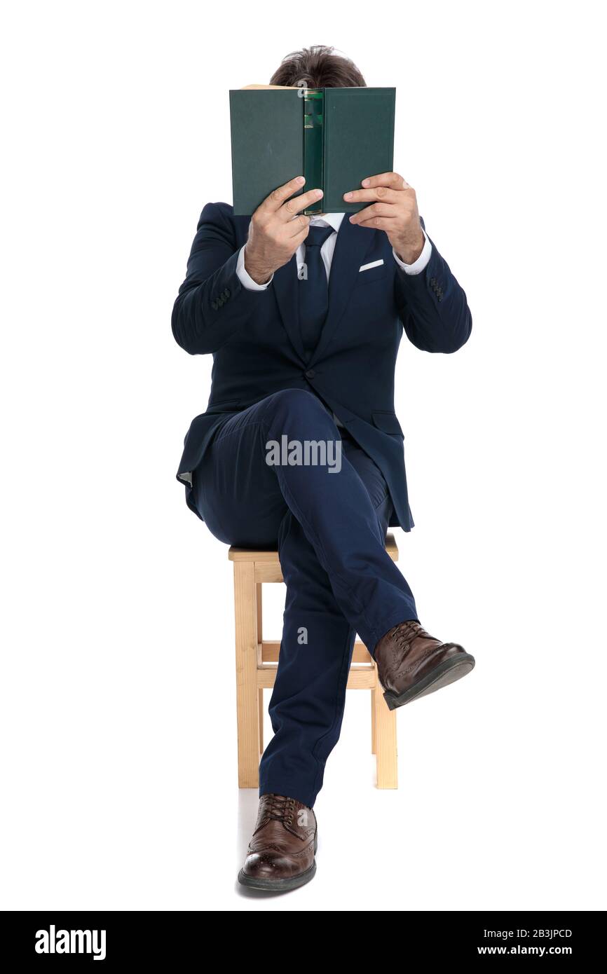 l'homme d'affaires formel avec costume de marine est assis avec des jambes croisées et le visage recouvert d'un livre sur fond de studio blanc Banque D'Images