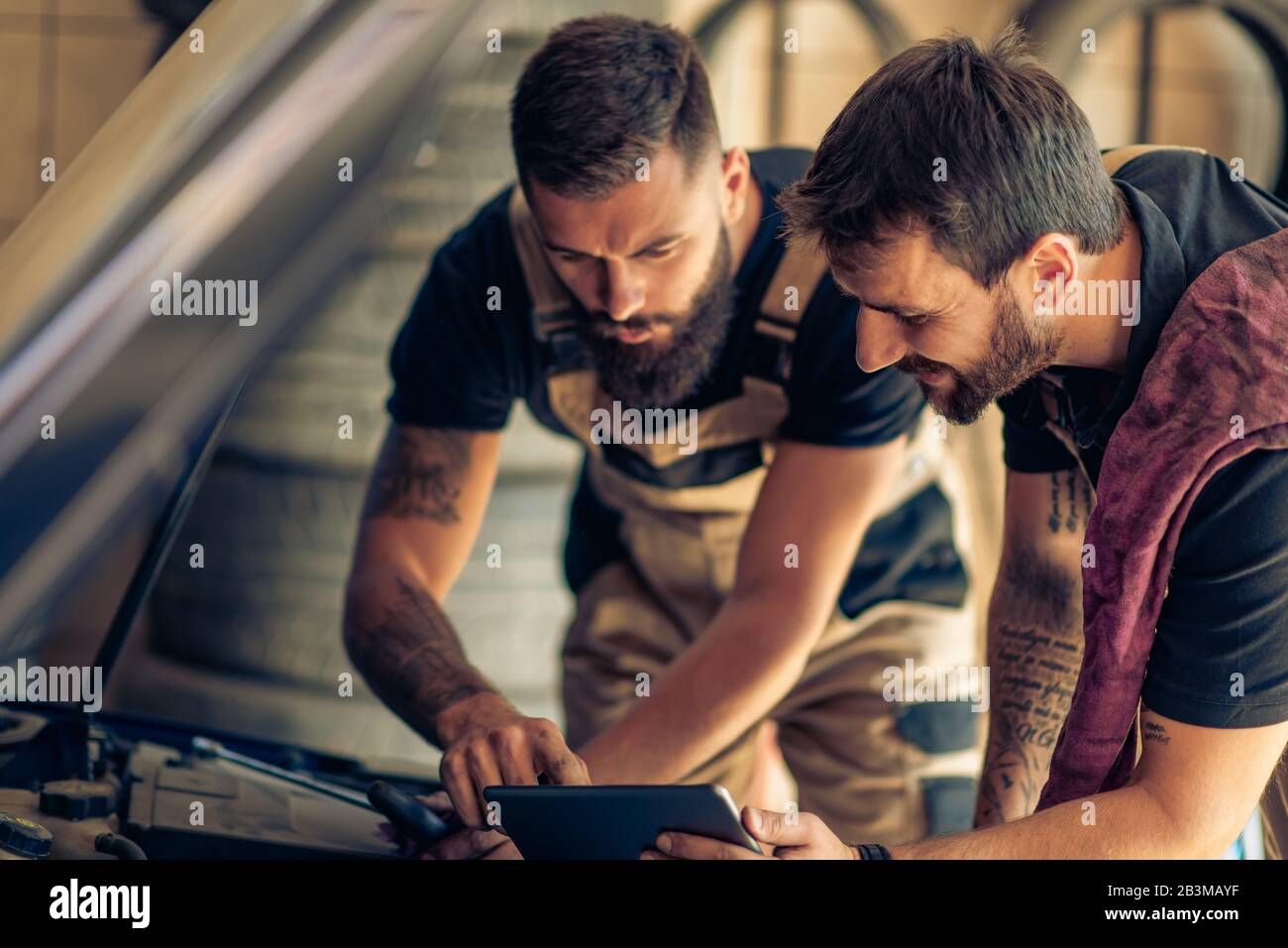 Deux mécaniciens travaillant sur des rapports dans un atelier.Mécanique avec outil d'acquisition diagnostiquant la voiture dans un capot ouvert. Banque D'Images