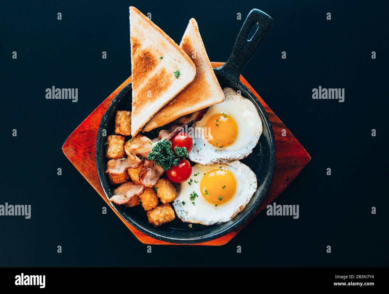 Cuisine de style brunch occidental avec toasts, œufs côté ensoleillé, tomates sur une plaque en bois et fond noir. Banque D'Images