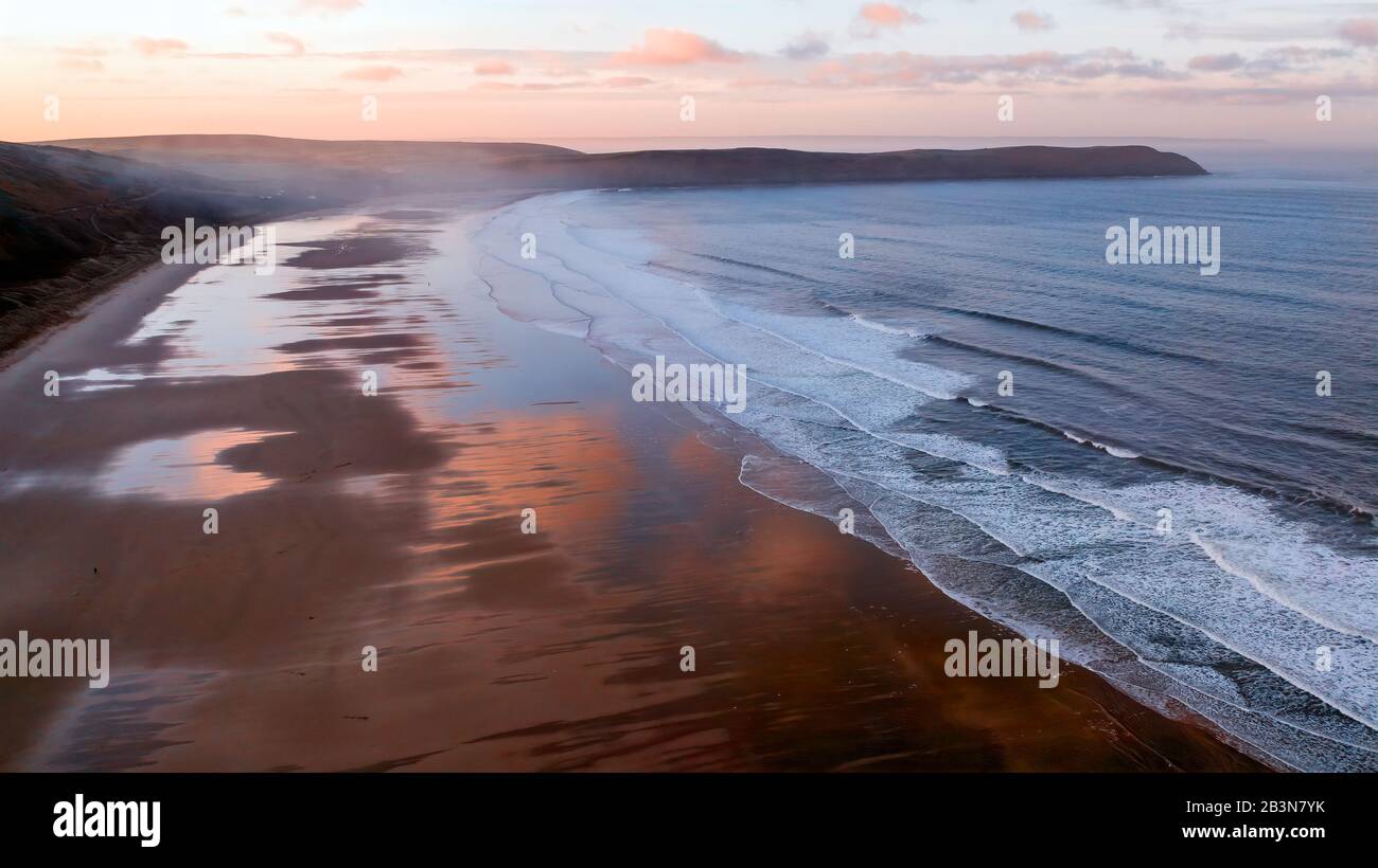 Vue aérienne de la plage de Woolacombe à l'aube, vagues se brisant sur une plage où les nuages éclairés par le soleil se reflètent dans l'eau debout. Headlands sur le raiz Banque D'Images