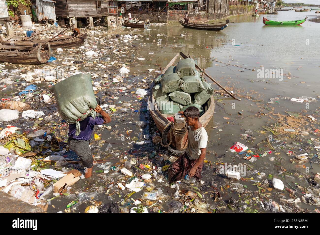 Deux hommes déchargeant des sacs de produits à partir d'un petit bateau dans le port de Sittwe, traversant une eau polluée pleine de déchets plastiques, Sittwe, Rakhine, Myanma Banque D'Images