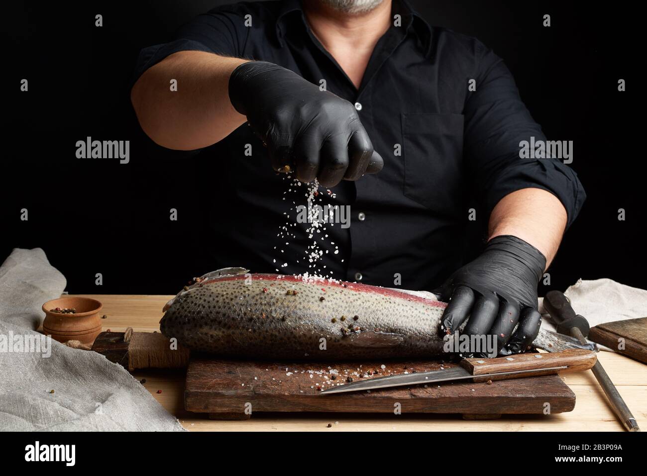 Chef dans une chemise noire et des gants en latex noir prépare un filet de  saumon sur planche à découper en bois, d'un processus d'aspersion avec les  épices et le sel et