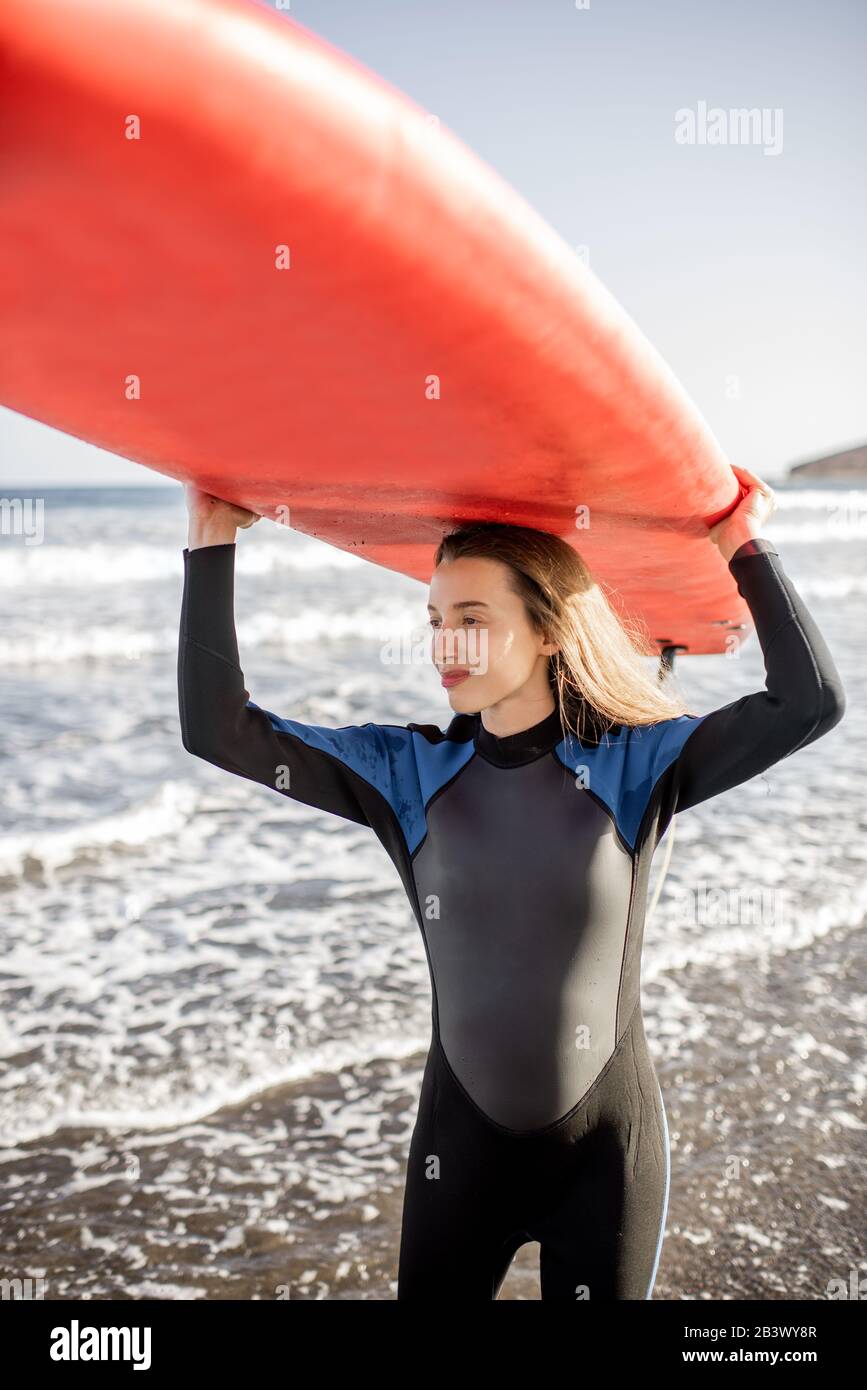 Portrait d'une jeune femme en combinaison portant planche de surf au-dessus  de la tête, se préparant à surfer sur la plage sauvage au coucher du  soleil. Concept de style de vie actif
