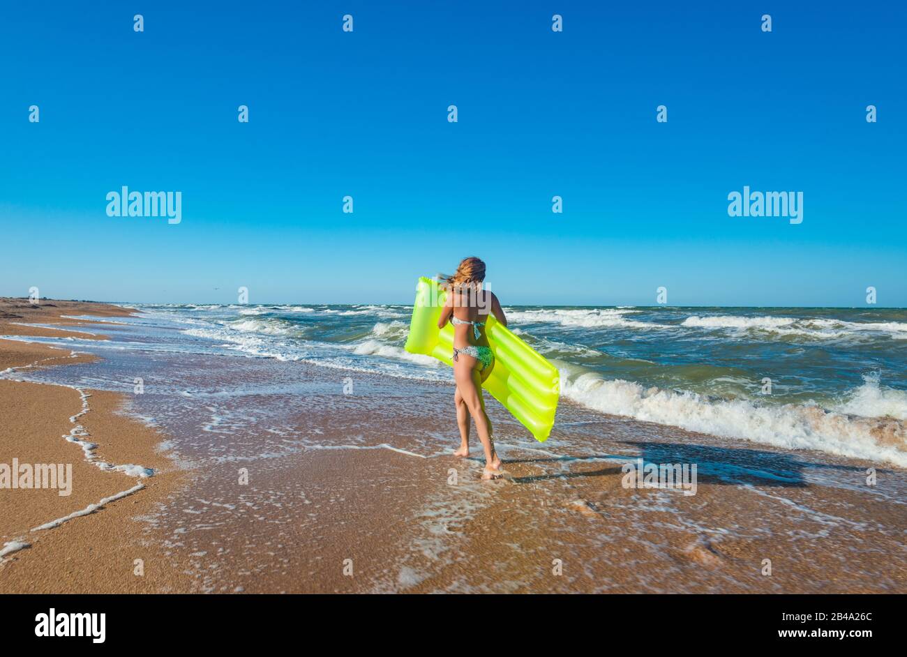 Belle jeune femme mince dans un maillot de bain court le long de la plage avec un matelas aérien le long de la rive sablonneuse près des vagues de la mer pluvieuse sur une somme chaude et ensoleillée Banque D'Images