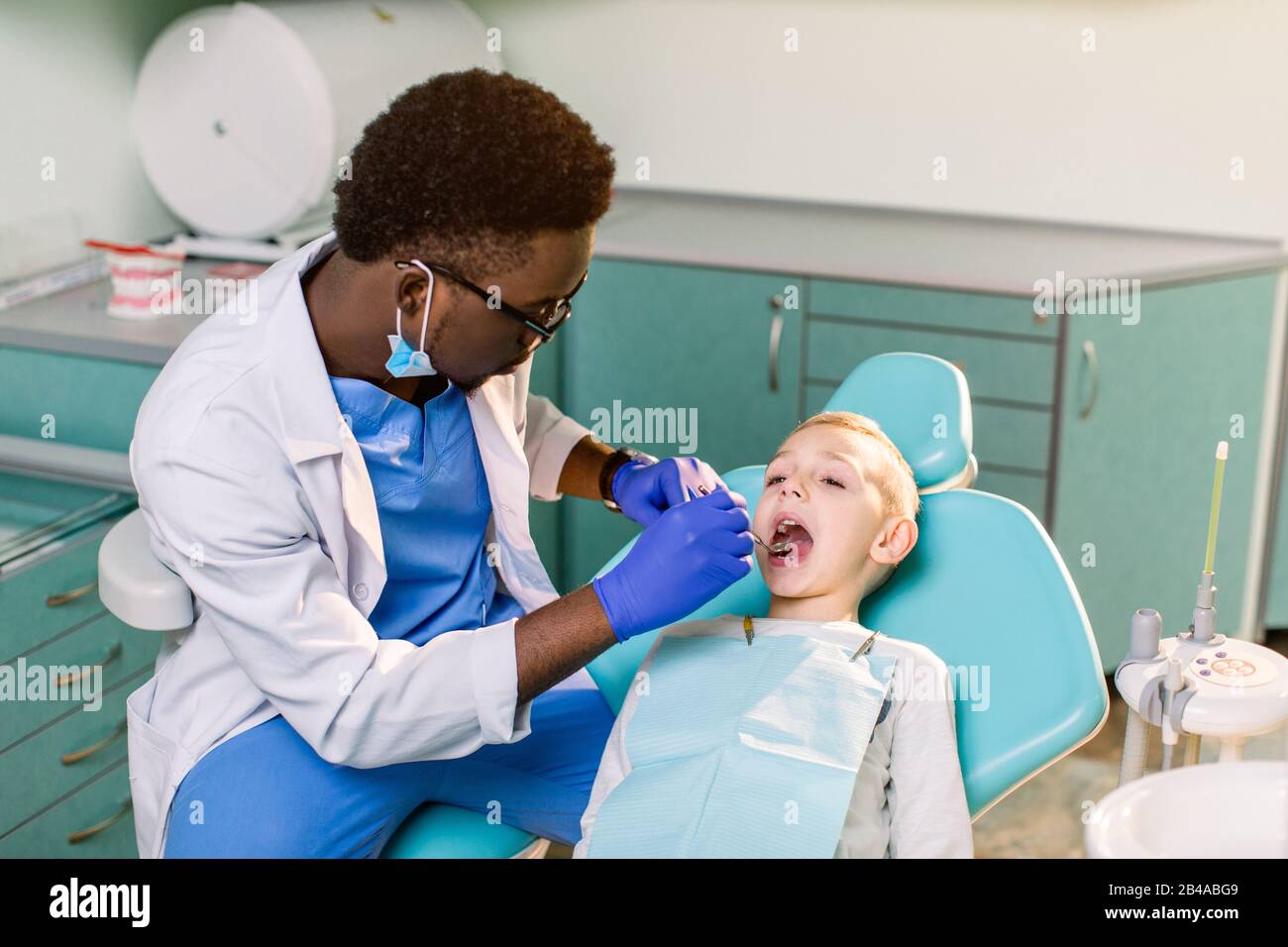 Dentiste masculin noir ethnique afro-américain avec gants en latex bleu  vérifiant l'état des dents d'un petit garçon souriant. Bébé garçon dans une  chaise dentaire bleue en mois Photo Stock - Alamy
