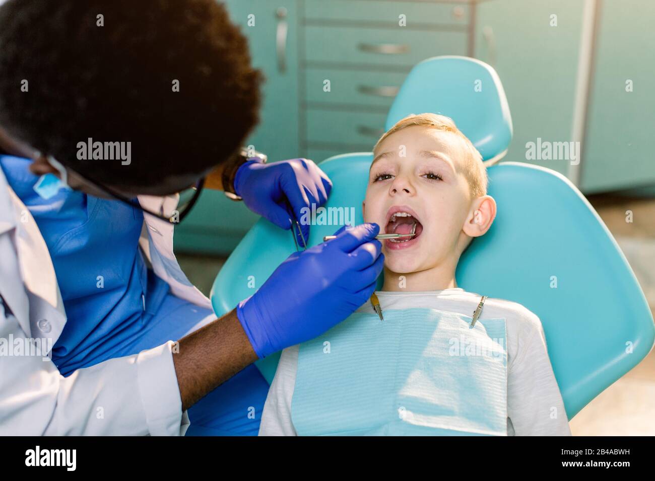 Dentiste Masculin Noir Ethnique Afro Americain Avec Gants En Latex Bleu Verifiant L Etat Des Dents D Un Petit Garcon Souriant Bebe Garcon Dans Une Chaise Dentaire Bleue En Mois Photo Stock Alamy